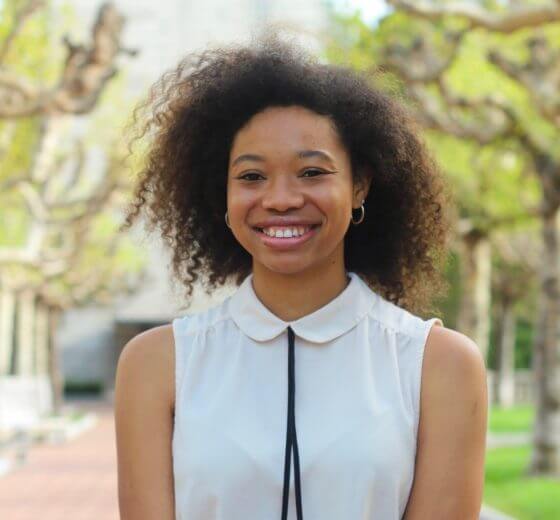 Headshot of miki oreggio smiling in front of trees. Wearing a white collared blouse with black tie in the middle.